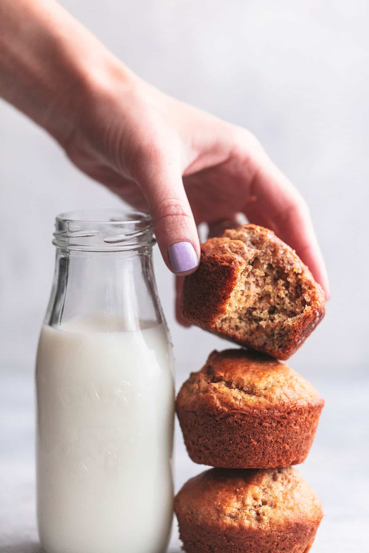 a hand picking up a bran muffin with a bite out stacked on top of more muffins with a bottle of milk on the side.