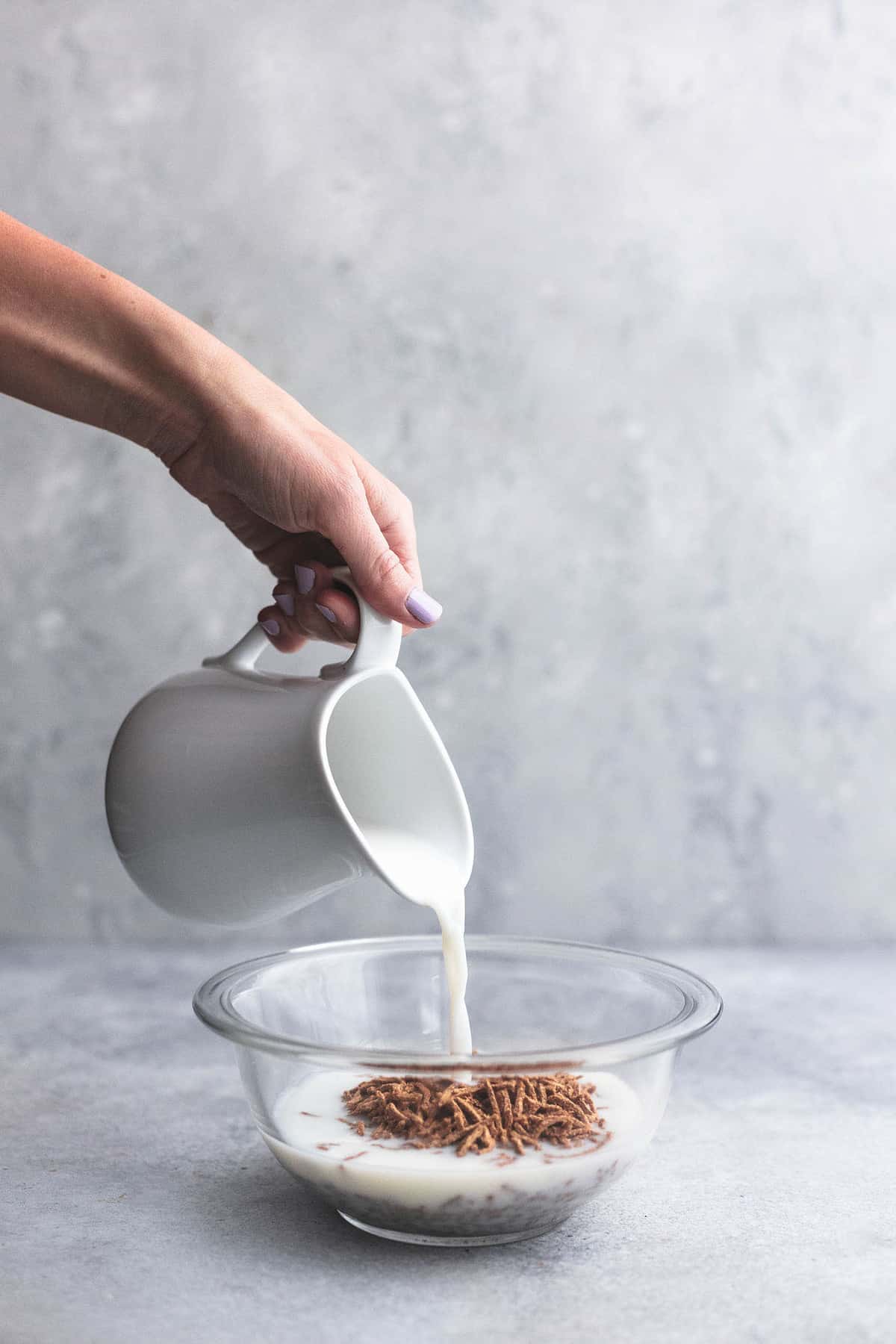 milk being poured from a white pitcher into a bowl of bran cereal in a glass bowl.