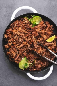 overhead view of tongs holding shredded pork with lime wedges in a cast iron pan