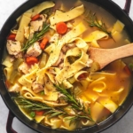 overhead view of serving spoon in pot of noodles with broth and chicken