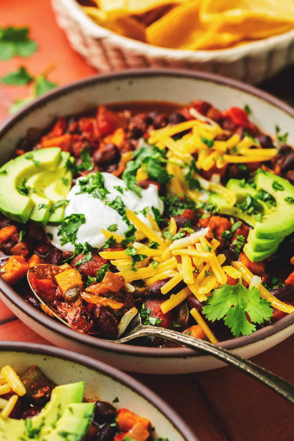 spoon scooping into bowl of chili topped with cheese, avocado, and sour cream