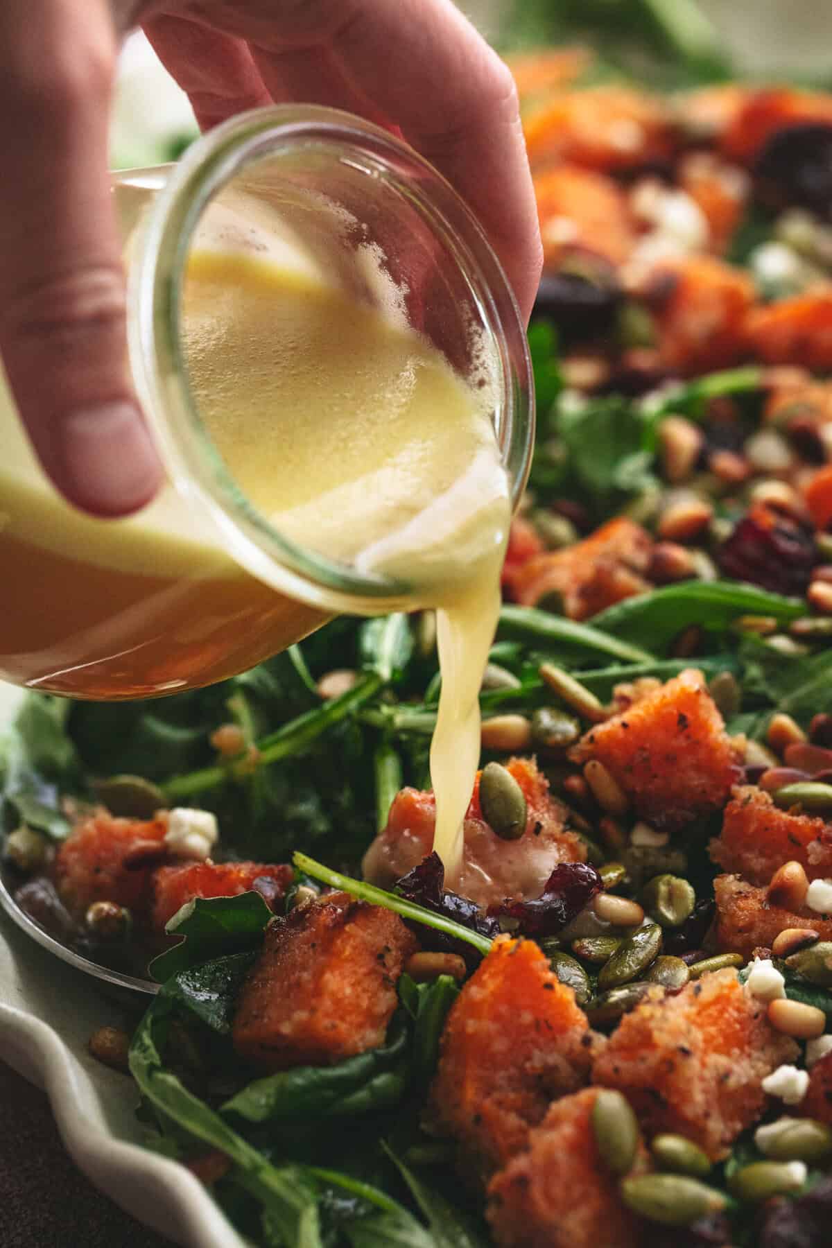 hand pouring cup of apple cider dressing onto butternut squash and arugula salad