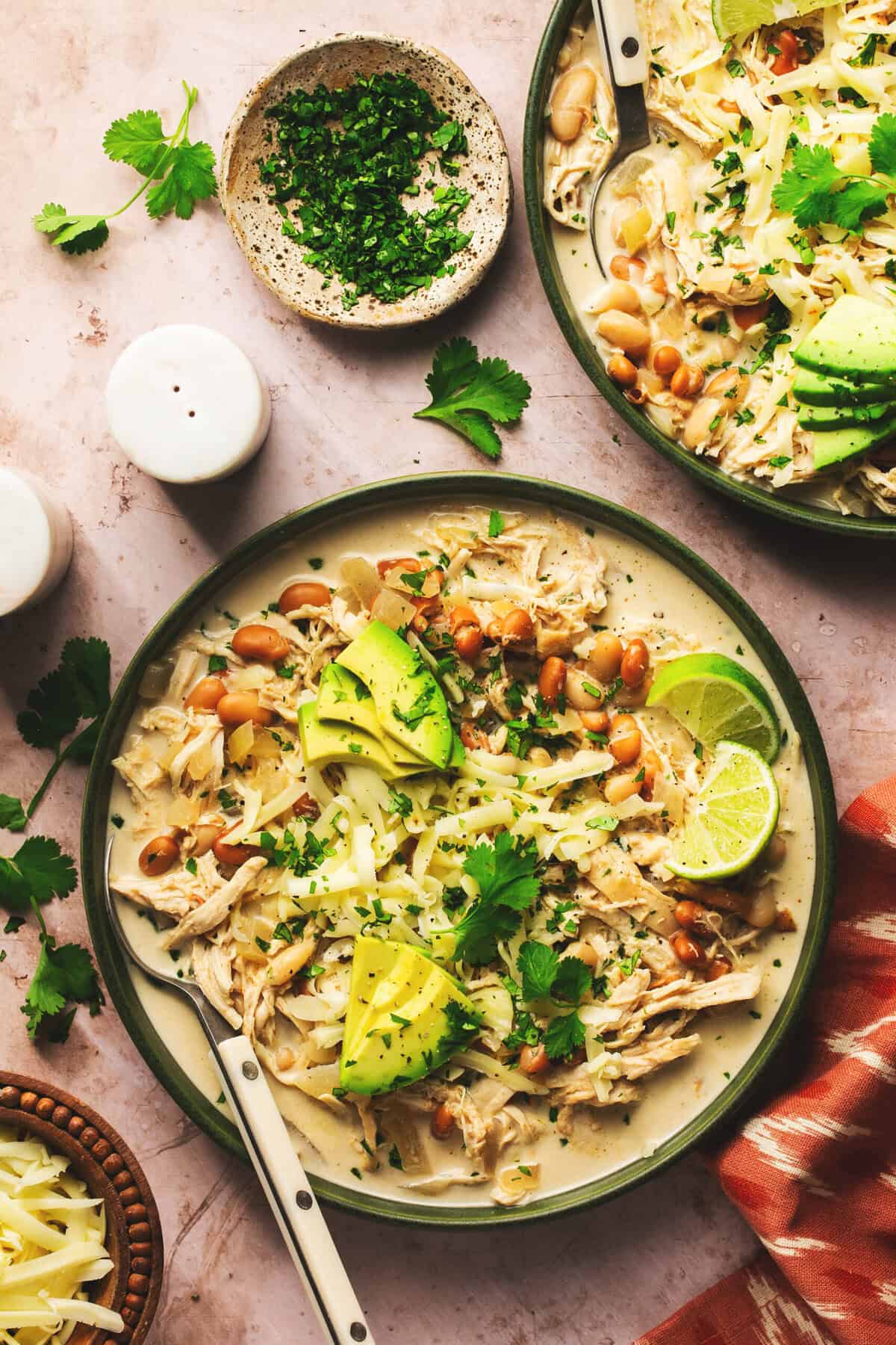 two bowls of slow cooker white chicken chili on table with garnishes and salt shaker